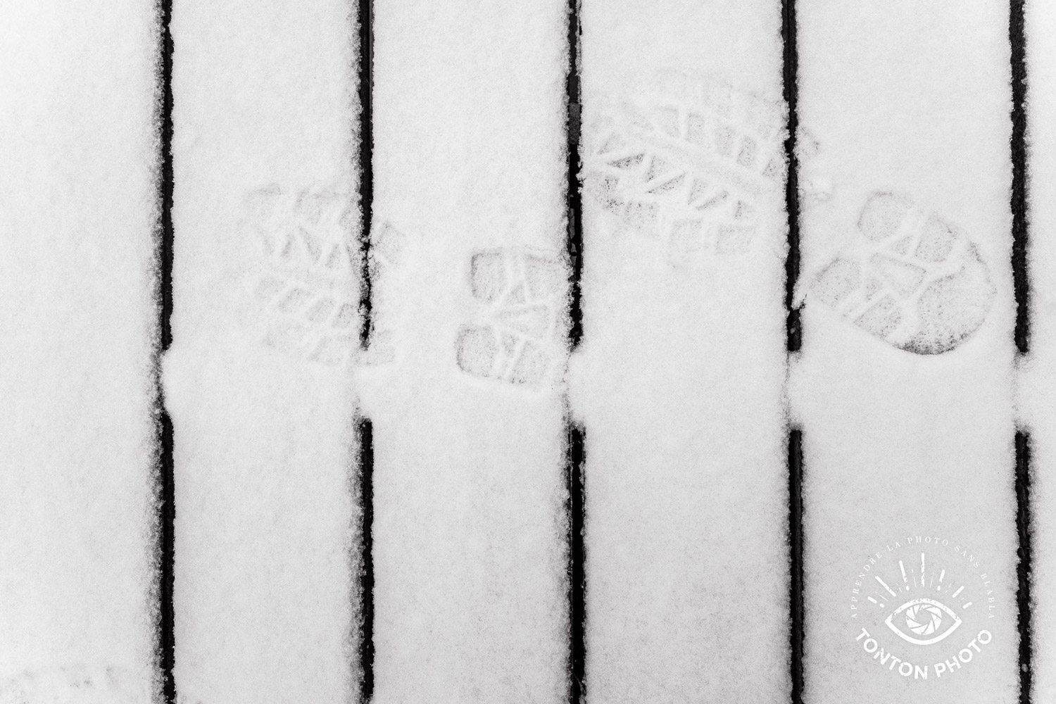 Traces de pas dans la neige sur une passerelle en bois, en ville. En observant et en soignant ma composition, j'ai joué sur la symétrie des lignes pour mettre en valeur ces traces © Tonton Photo